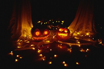 Two carved pumpkins with glowing faces are positioned in a dimly lit setting. They are surrounded by a string of yellow fairy lights, creating a warm and festive ambiance. White fabric drapes are in the background, enhancing the Halloween-themed atmosphere.