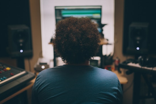 A person with curly hair sits in front of a computer monitor in a music studio. Surrounding the individual are various pieces of audio equipment, including speakers and a mixing console. Soft lighting and a slightly blurred background suggest a focused work environment.