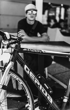 A black and white image features a bicycle with prominent branding, positioned in the foreground. In the background, a person wearing a cap and sunglasses is seated at a table, with folded arms, slightly out of focus.