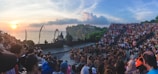 A large crowd gathers in an amphitheater to watch a traditional Balinese Kecak dance performance at sunset. Performers are seated in a circular formation, surrounded by ocean views and dramatic cliffs.