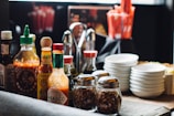 A collection of various sauces, spices, and condiments arranged on a table. There are multiple bottles of hot sauce, including brands like Tabasco and Sriracha, alongside jars of crushed red pepper and utensils. Stacks of small white plates are also visible, adding to the organized appearance of the dining setup.