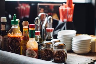 A collection of various sauces, spices, and condiments arranged on a table. There are multiple bottles of hot sauce, including brands like Tabasco and Sriracha, alongside jars of crushed red pepper and utensils. Stacks of small white plates are also visible, adding to the organized appearance of the dining setup.