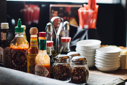 A collection of various sauces, spices, and condiments arranged on a table. There are multiple bottles of hot sauce, including brands like Tabasco and Sriracha, alongside jars of crushed red pepper and utensils. Stacks of small white plates are also visible, adding to the organized appearance of the dining setup.