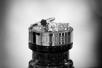 A pair of elegant rings are stacked atop the metallic cap of a bottle, which is engraved with the word 'CLUB'. The rings feature prominent gemstones and intricate detailing, with one ring showcasing a large circular stone surrounded by smaller gems.