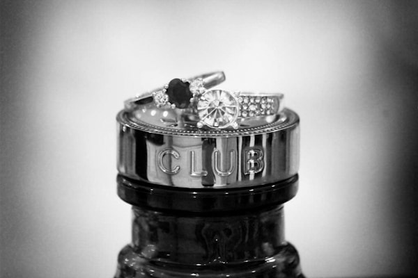 A pair of elegant rings are stacked atop the metallic cap of a bottle, which is engraved with the word 'CLUB'. The rings feature prominent gemstones and intricate detailing, with one ring showcasing a large circular stone surrounded by smaller gems.