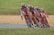 A group of cyclists dressed in matching red and white uniforms is racing closely together on a road. They are leaning into a turn, showcasing teamwork and speed. The background features a grassy area and a section of the racetrack.