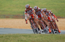 A group of cyclists dressed in matching red and white uniforms is racing closely together on a road. They are leaning into a turn, showcasing teamwork and speed. The background features a grassy area and a section of the racetrack.