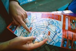Two hands are holding and pointing at a colorful city map. The map contains various streets, landmarks, and highlighted routes. Important points are circled in blue, with sections of the map bordered in red.