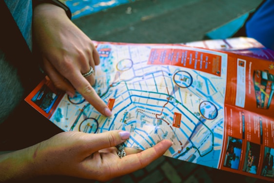 Two hands are holding and pointing at a colorful city map. The map contains various streets, landmarks, and highlighted routes. Important points are circled in blue, with sections of the map bordered in red.
