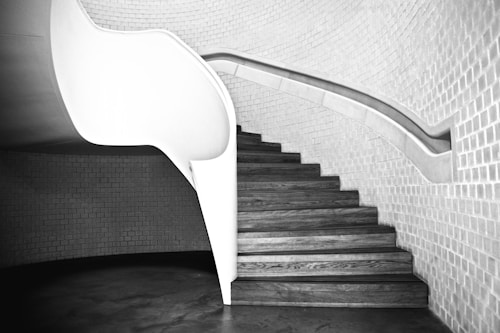 A curved staircase with wooden steps and a sleek, white balustrade is set against a backdrop of smooth, light-colored brick walls. The design is modern and minimalistic, emphasizing clean lines and the contrast between light and shadow.