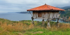 A rustic wooden granary stands on stone pillars with a red tiled roof, set against a coastal backdrop. The sea is visible in the distance under an overcast sky, and the surrounding landscape is lush with grass and cliffs.