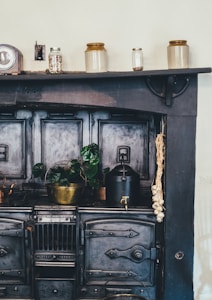 A vintage kitchen stove is adorned with pots and jars. There are three ceramic jars and a smaller glass jar placed on a narrow shelf above the stove. A brass pot with a leafy green plant sits on top of the stove, alongside a black kettle. A bundle of garlic hangs from the side, adding a rustic touch.