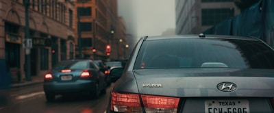 A street scene featuring several cars, including a Hyundai with a Texas license plate in the foreground. The road is lined with tall buildings and enveloped in fog, creating a moody atmosphere. The taillights and traffic lights provide spots of color against the muted tones.