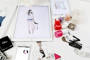 A workspace is filled with feminine fashion illustrations and cosmetic items. Digital drawings are displayed on tablets alongside traditional sketches. Nail polish bottles in various vibrant colors and a Dior makeup box are arranged neatly. The scene blends technology and art in a stylish manner.