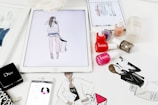A workspace is filled with feminine fashion illustrations and cosmetic items. Digital drawings are displayed on tablets alongside traditional sketches. Nail polish bottles in various vibrant colors and a Dior makeup box are arranged neatly. The scene blends technology and art in a stylish manner.