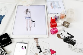 A workspace is filled with feminine fashion illustrations and cosmetic items. Digital drawings are displayed on tablets alongside traditional sketches. Nail polish bottles in various vibrant colors and a Dior makeup box are arranged neatly. The scene blends technology and art in a stylish manner.
