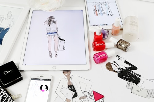 A workspace is filled with feminine fashion illustrations and cosmetic items. Digital drawings are displayed on tablets alongside traditional sketches. Nail polish bottles in various vibrant colors and a Dior makeup box are arranged neatly. The scene blends technology and art in a stylish manner.