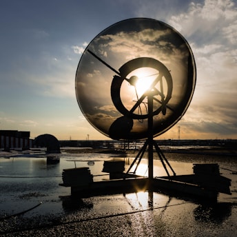 A large satellite dish silhouetted against a setting sun. The sun's rays create a striking effect through the perforated surface of the dish. The foreground is dark and reflective due to water on the ground, while scattered clouds fill the background sky.