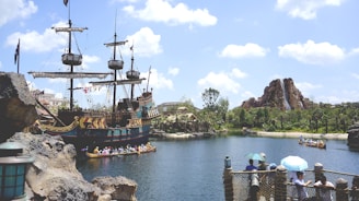 A large pirate ship with masts and sails is docked near rocky terrain beside a serene body of water. People in colorful outfits are paddling a canoe on the water, and a path surrounded by foliage leads to a mountainous structure with a waterfall. A few individuals are observing from a wooden bridge with lanterns, and the sky is bright with clouds.