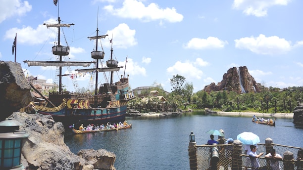 A large pirate ship with masts and sails is docked near rocky terrain beside a serene body of water. People in colorful outfits are paddling a canoe on the water, and a path surrounded by foliage leads to a mountainous structure with a waterfall. A few individuals are observing from a wooden bridge with lanterns, and the sky is bright with clouds.