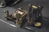 Two traditional coil tattoo machines are placed on a dark surface, accompanied by tattoo needles. The machines are made of brass and metal components, showcasing a vintage design.