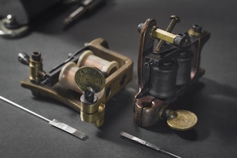 Two traditional coil tattoo machines are placed on a dark surface, accompanied by tattoo needles. The machines are made of brass and metal components, showcasing a vintage design.