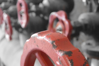 A close-up view of several industrial red valve handles, with a blurred background. The metal is weathered with visible signs of rust and wear.