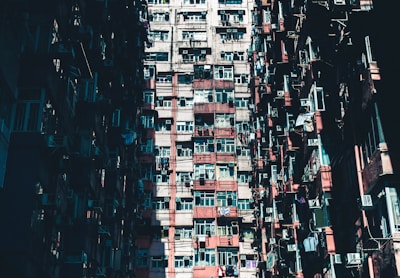 A densely packed residential building with numerous windows and balconies, displaying a mix of air conditioning units and laundry hanging outside. The structure has a mix of red and beige tones, with parts of the façade showing signs of weathering.