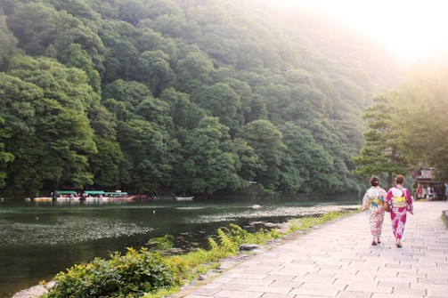 Two people walk along a stone path beside a serene river, with lush green trees covering the hills in the background and boats gently floating on the water. The scene is peaceful and sunlit, creating a harmonious blend of nature and tradition.