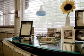 The image features a table by a window with several personal and decorative items. A framed document with deer antlers, several framed photographs, and some decorative wooden blocks are present. A large baby bottle and a bright sunflower in a vase add to the scene, with blinds partially covering the window.