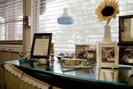 The image features a table by a window with several personal and decorative items. A framed document with deer antlers, several framed photographs, and some decorative wooden blocks are present. A large baby bottle and a bright sunflower in a vase add to the scene, with blinds partially covering the window.