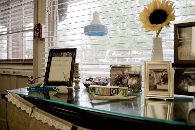 The image features a table by a window with several personal and decorative items. A framed document with deer antlers, several framed photographs, and some decorative wooden blocks are present. A large baby bottle and a bright sunflower in a vase add to the scene, with blinds partially covering the window.