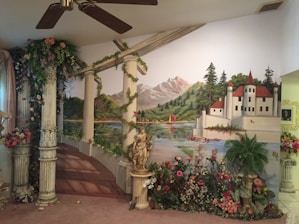 A detailed mural on a wall depicting a picturesque landscape with a lake, surrounded by mountains and forests, and a white castle with red roofs. In the foreground, there are classical Greek-style columns adorned with flowers and ivy, along with a sculpture, and a variety of colorful flowers and plants.
