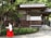 A person dressed in traditional Japanese attire walks along a pathway in front of a wooden building surrounded by lush greenery. The structure features wooden beams and white walls, set against a backdrop of tall trees.