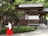 A person dressed in traditional Japanese attire walks along a pathway in front of a wooden building surrounded by lush greenery. The structure features wooden beams and white walls, set against a backdrop of tall trees.