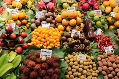 A vibrant display of various fruits and colorful produce, neatly arranged with labels indicating their names and origins. The assortment includes starfruits, naranjillas, rambutans, passion fruits, and more, accompanied by a bottle of juice or syrup. The fruits are placed on green leafy backdrops, enhancing their freshness and appeal.