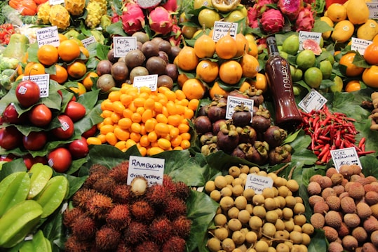 A vibrant display of various fruits and colorful produce, neatly arranged with labels indicating their names and origins. The assortment includes starfruits, naranjillas, rambutans, passion fruits, and more, accompanied by a bottle of juice or syrup. The fruits are placed on green leafy backdrops, enhancing their freshness and appeal.