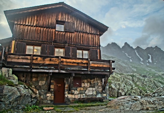 A rustic, wooden mountain cabin is situated amidst rocky terrain with a backdrop of jagged, snow-capped peaks under a partly cloudy sky. The cabin's lower section is made of stone, and there are visible wooden shutters and a small porch area.