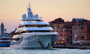 A large, modern yacht is anchored near a historic waterfront with colorful, old buildings in the background. The setting is during sunset, casting a warm glow over the scene. The yacht features multiple decks and intricate design details.