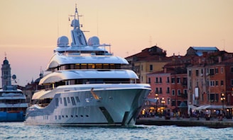 A large, modern yacht is anchored near a historic waterfront with colorful, old buildings in the background. The setting is during sunset, casting a warm glow over the scene. The yacht features multiple decks and intricate design details.
