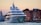 A large, modern yacht is anchored near a historic waterfront with colorful, old buildings in the background. The setting is during sunset, casting a warm glow over the scene. The yacht features multiple decks and intricate design details.