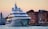 A large, modern yacht is anchored near a historic waterfront with colorful, old buildings in the background. The setting is during sunset, casting a warm glow over the scene. The yacht features multiple decks and intricate design details.