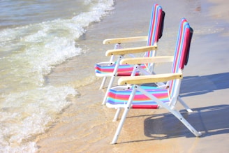 Two colorful striped beach chairs are positioned on the sandy shore, facing the gentle waves of the ocean. The light from the sun casts a warm glow on the sand and water, contributing to a relaxing and serene atmosphere.