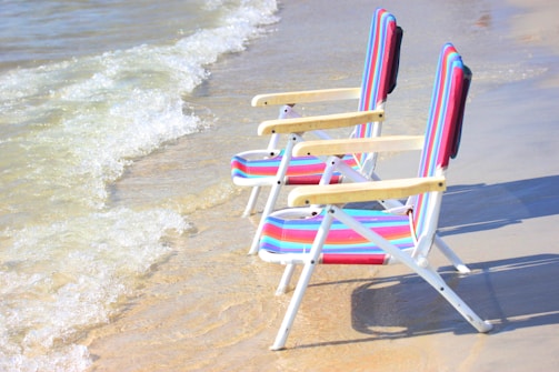 Two colorful striped beach chairs are positioned on the sandy shore, facing the gentle waves of the ocean. The light from the sun casts a warm glow on the sand and water, contributing to a relaxing and serene atmosphere.