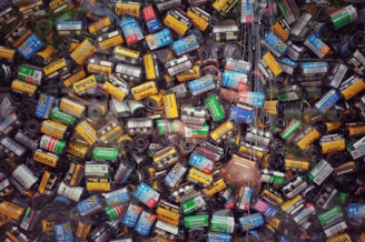 A large collection of colorful film canisters is densely arranged, with labels like Kodak, Fujicolor, and others visible on the spools. The canisters are in various colors, including yellow, red, blue, green, and black, creating a vibrant mosaic.