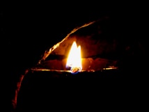 A single flame burns brightly in the darkness, providing a warm and gentle glow. The flame is surrounded by a pitch-black background, emphasizing its light and energy.