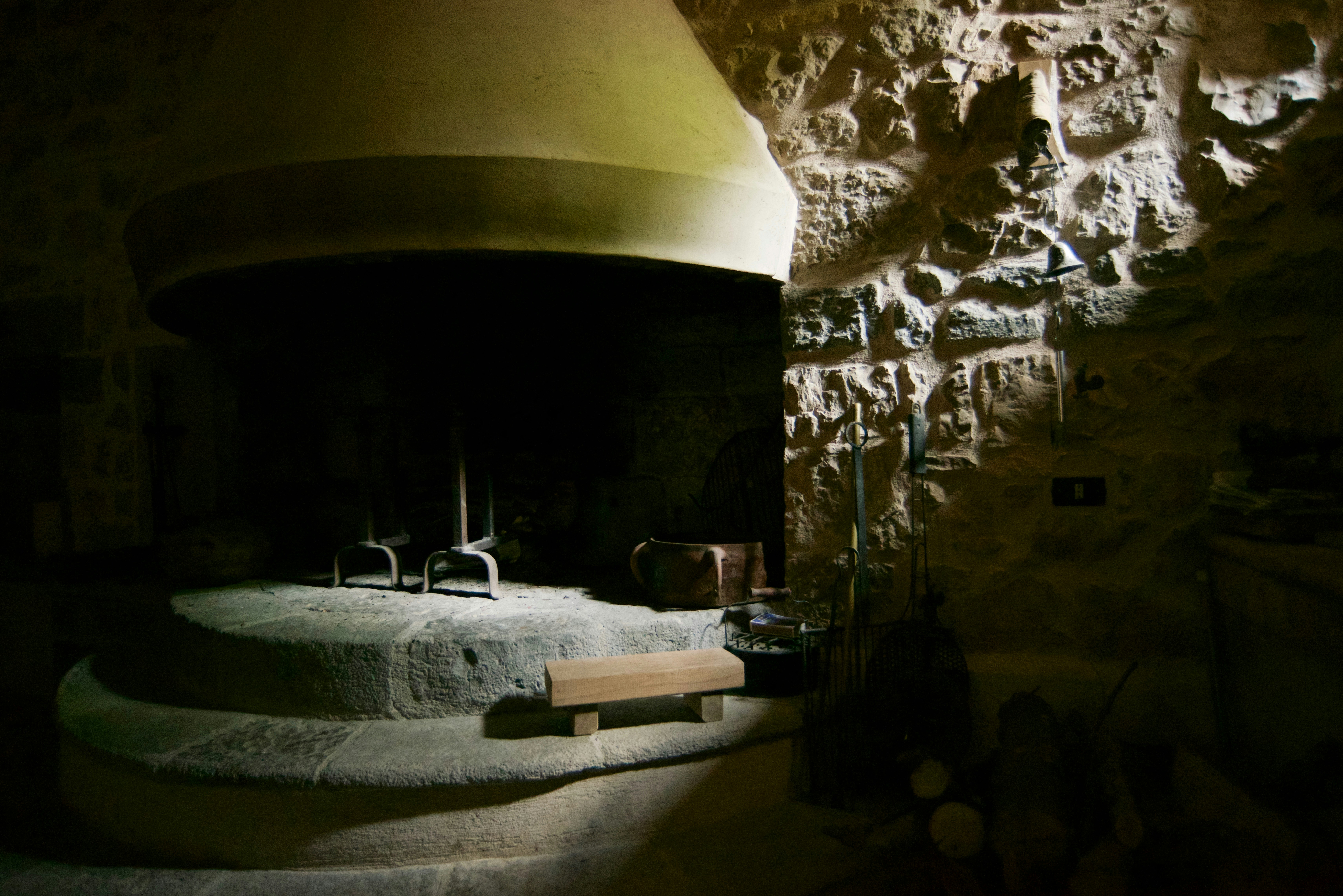 A rustic interior space with a large, old-fashioned stone fireplace set against a textured stone wall. The lighting is dim and atmospheric, casting shadows on the wall and highlighting the rough texture of the stone. There are various objects near the fireplace, including a metal stand, a small wooden bench, and a few rustic tools.