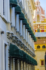 A facade of a building with multiple green canopies aligned in a row, featuring detailed architectural elements in a classic style. In the background, an ornate dome with pink and yellow details rises, adding a vibrant contrast to the scene.