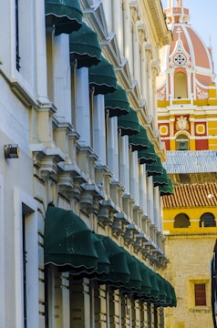 A facade of a building with multiple green canopies aligned in a row, featuring detailed architectural elements in a classic style. In the background, an ornate dome with pink and yellow details rises, adding a vibrant contrast to the scene.
