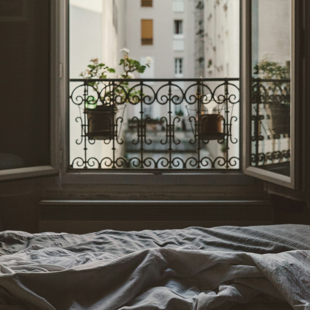 An open window at the foot of an bed with messy sheets looks at flowers on the sill and other buildings across the way